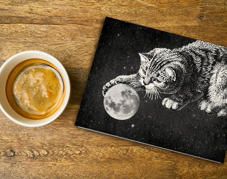 A small cup of espresso is next to a black card on a wood table. The card has a cat playing with a full moon. Both are realistic images in black and white.