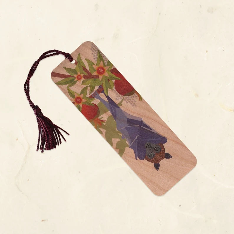 A wood bookmark with a burgundy tassel. It has a purple, gray, and brown fruit bat hanging from a berry plant.