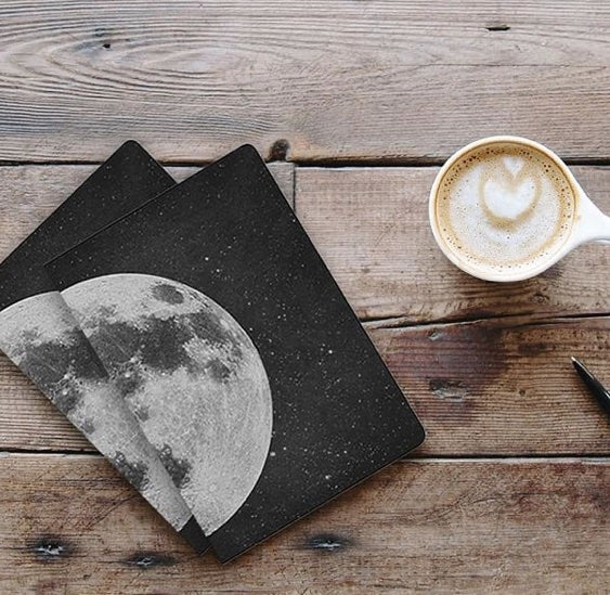 Two notebooks on a wood table. They are black with a large image of the moon on the left side. One is stacked on top of the other and there is a small cup of espresso to the right.