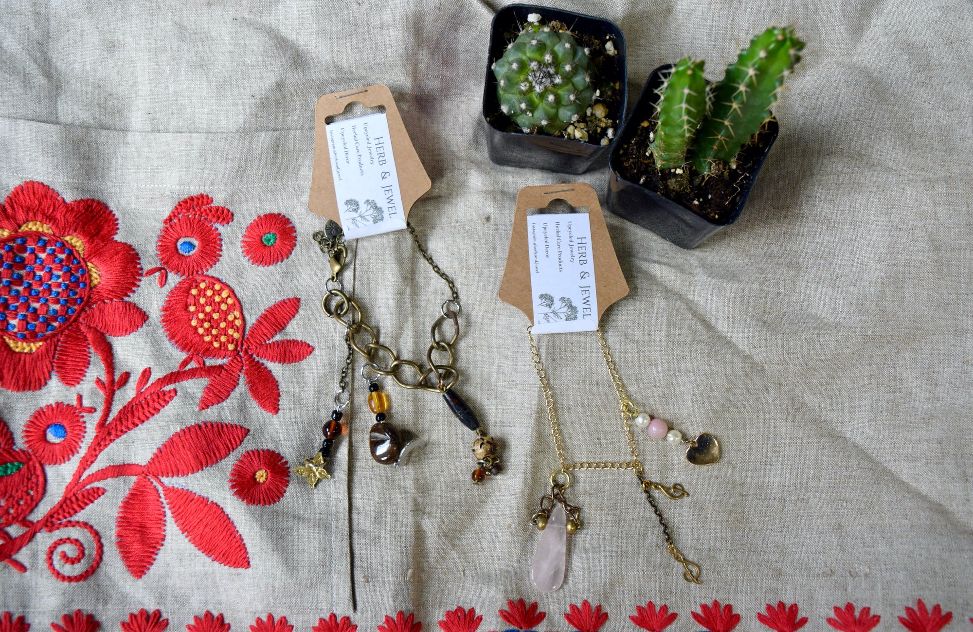 Two bracelets next to two small green cacti. The first bracelet has a dark golden chain with brown hanging beads and charms. The second has a light gold chain with golden hanging charms and pink and white beads or gems.