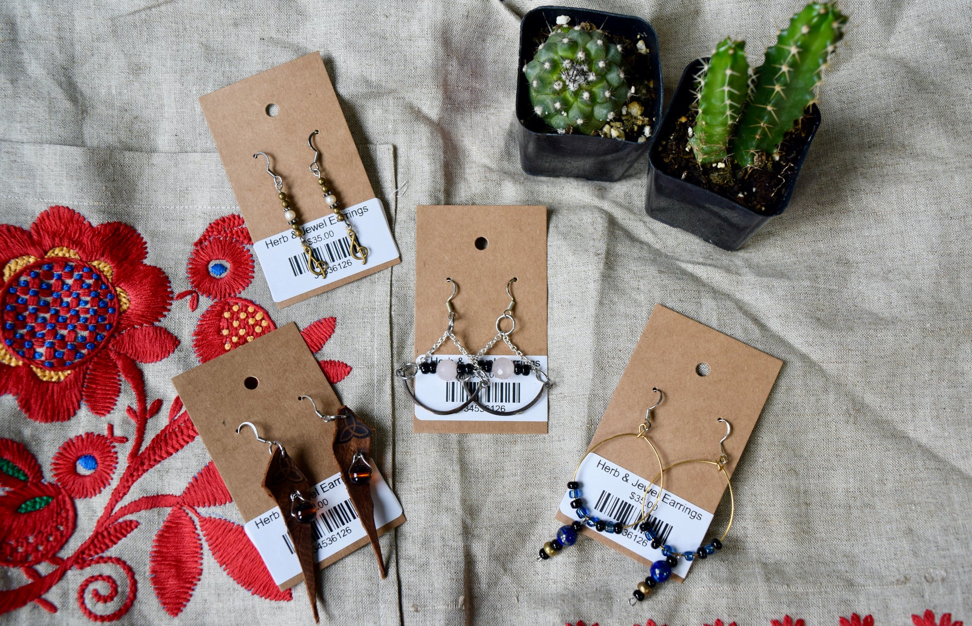 Several different pairs of earrings next to 2 small green cacti. The earrings range from hanging to hoops and all have beads of varying colors. Each has gold or silver metal.