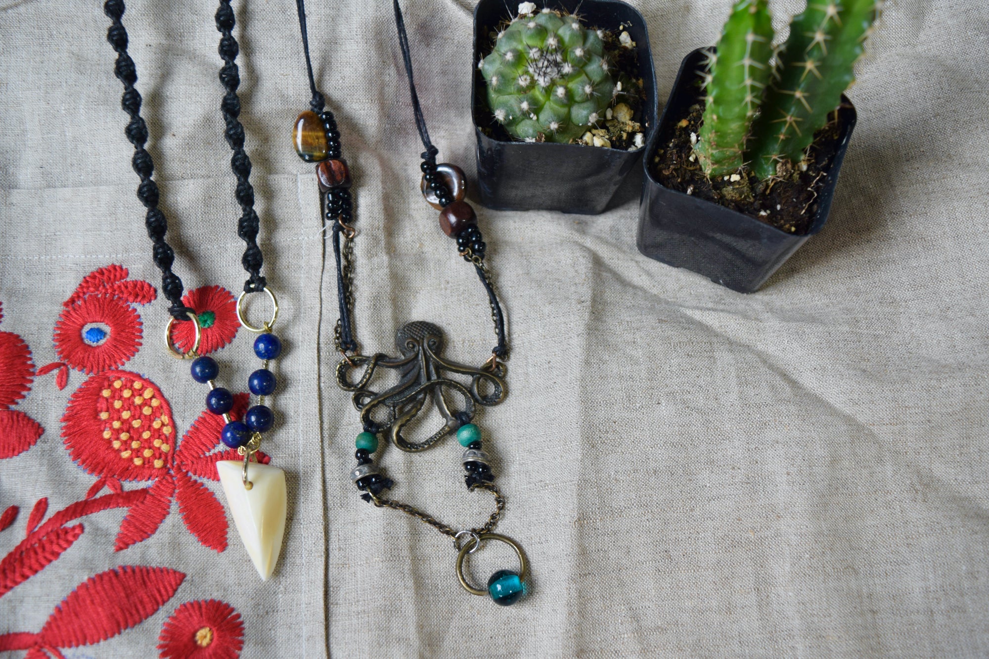 Two necklaces next to two small green cacti. The first necklace has a twisted black fiber chain with blue beads and a white stone hanging from it. The other has a thin black fiber chain with tiger's eye beads. Towards the bottom there is a dark metal octopus. Below it there are beads of various colors.