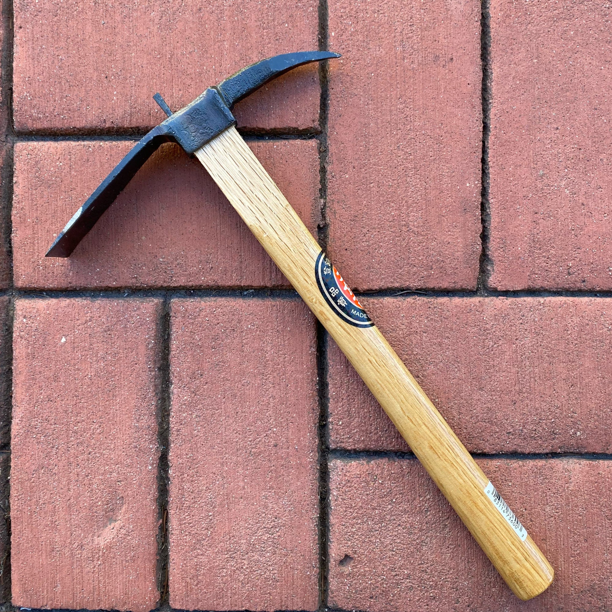 A tool with a black metal pick and a wooden handle. It is sitting on top of red bricks.