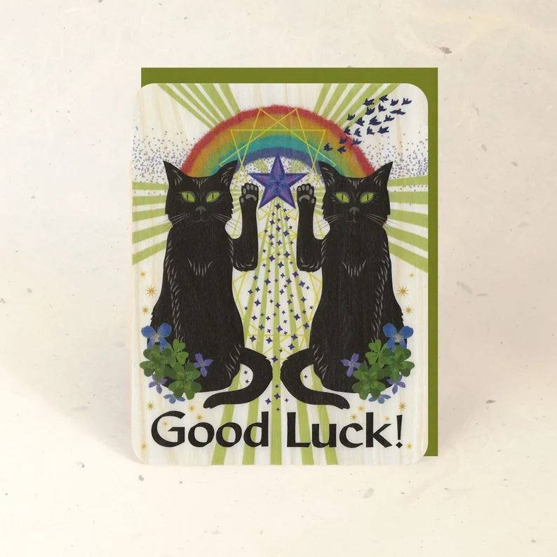 Two black cats sitting side by side with one paw raised. There is a rainbow and other designs in the center with green rays radiating outwards. The card says "Good Luck" at the bottom.
