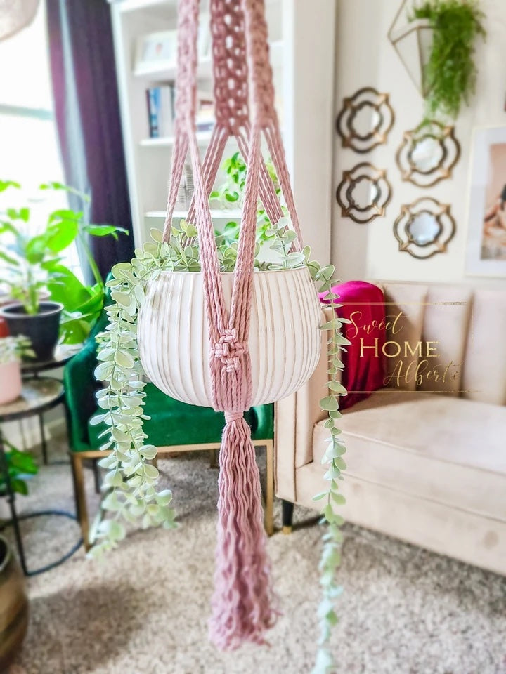 A rose macrame hanger with an intricate design. It is holding a white pot with a green hanging plant.