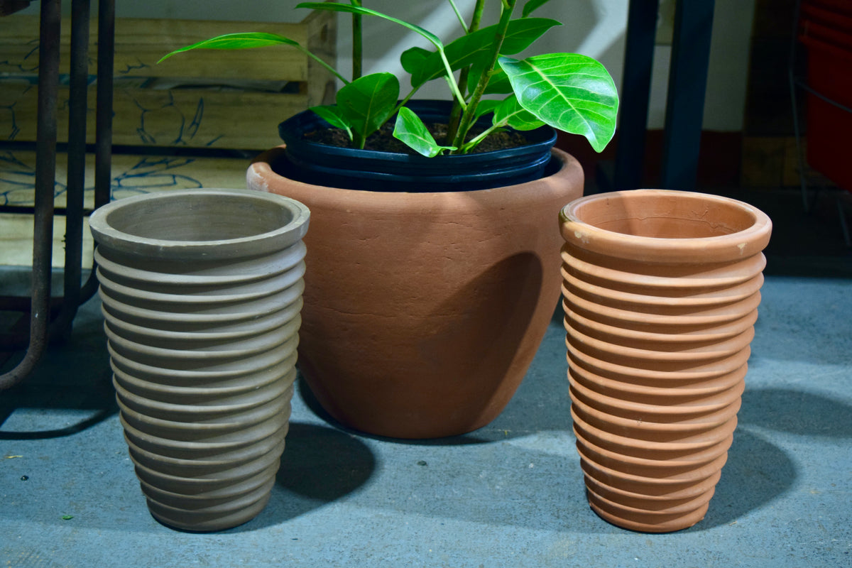 Two long, thin pots with horizontal ridges along the sides. One is gray and the other is terracotta. There is a large potted plant in the background.