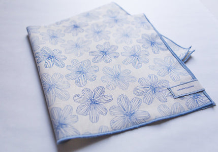 A folded white piece of cloth with a light blue boarder and print of blue flax flowers.