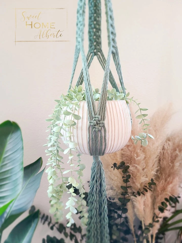 A mint green macrame hanger with an intricate design. It is holding a white pot with a green hanging plant.