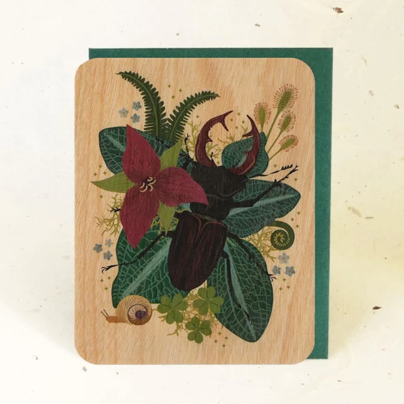 A card with a wooden background and green envelop featuring a black stag beetle surrounded by leaves, ferns, and flowers.