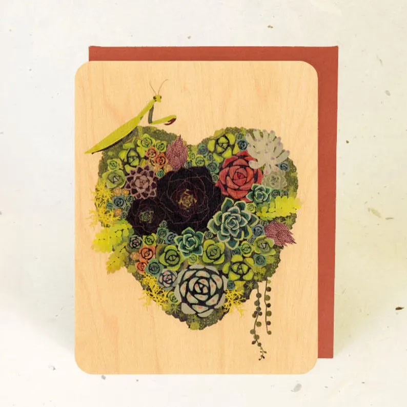 A wooden card with a red envelop featuring a heart made of various succulents. The succulents are many different colors, ranging from green to red. There is a lime green praying mantis on the top left corner of the heart.