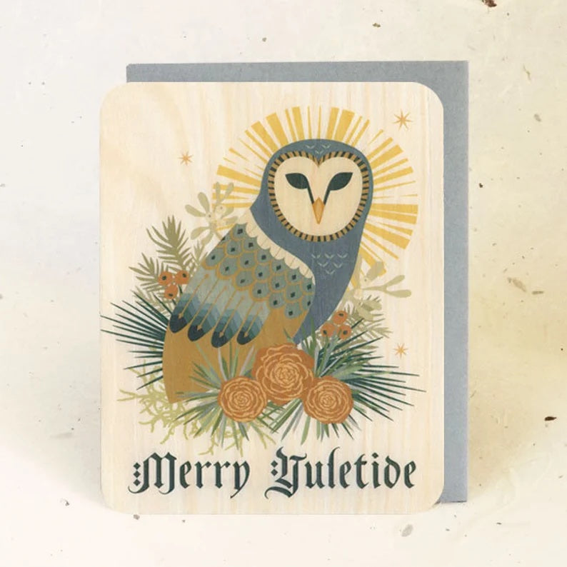 A wooden card with a barn owl sitting atop pine branches with pinecones. It reads "Merry Yuletide" at the bottom.