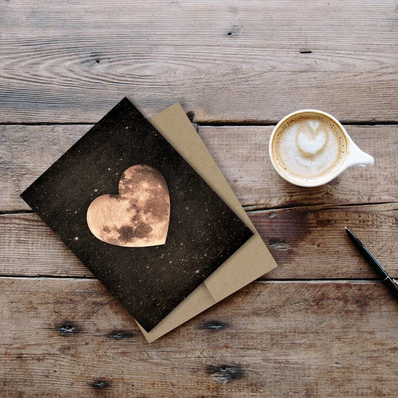 A black card with a starry background on top of a brown envelop and wooden table. The card has a brown heart shaped moon in the center. There is a small white espresso and black pen on the right.